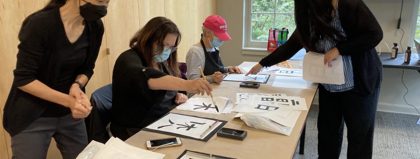 Several people working on calligraphy while sitting at a table while the instructor looks over their shoulders