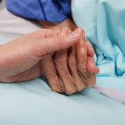 Young hand holding older, wrinkled hand on top of hospital blanket