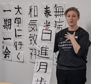 Girl standing with strip of calligraphy in front of a board covered in Japanese calligraphy