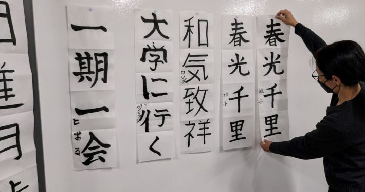 Vertical strips of Japanese calligraphy being hung on whiteboard by a teacher
